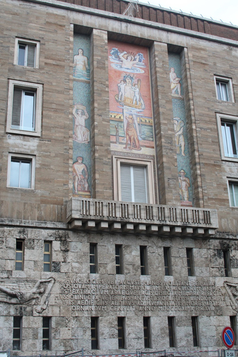 A photo of a side of a building with text engravings and angel figures on it. Also above a balcony there are mosaic pictures showing ancient people doing different jobs.