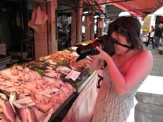 A woman taking a photo of some seafood stands.