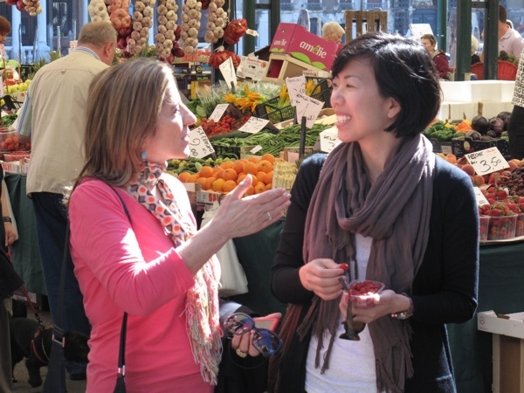 A photo of two women talking while standing near a fruit market.