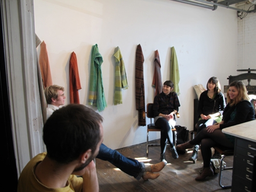 A photo of a group of people sitting in a classroom and talking while on the wall there ae some colorful scarfs hanged.