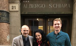 A photo of three people standing in front of a 3 star hotel that has the text: 21 ALBERGO S. CHIARA on it.