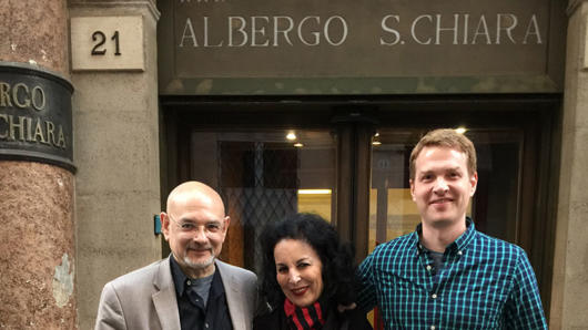 A photo of three people standing in front of a 3 star hotel that has the text: 21 ALBERGO S. CHIARA on it.