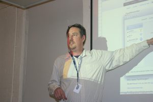 A photo of a man in a white shirt giving a lecture in front of a projector screen.