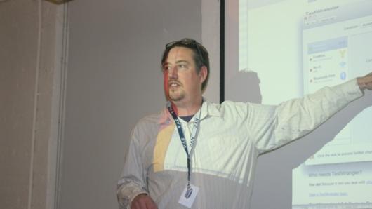 A photo of a man in a white shirt giving a lecture in front of a projector screen.