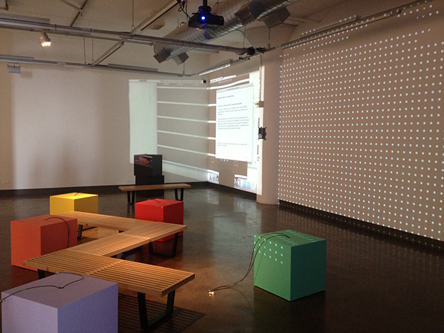 A photo of an art gallery setup with projected walls, wood benches and some colorful cubical shaped tables.