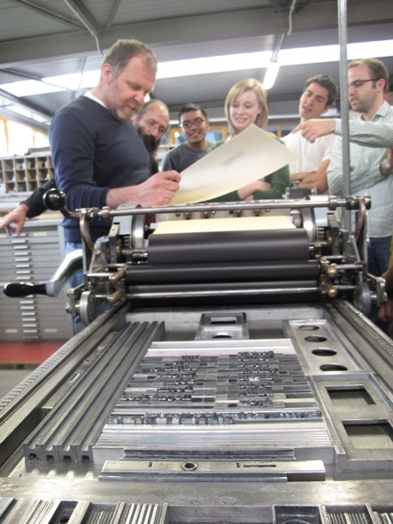 A photo of a group of people checking a paper from a typography machine.