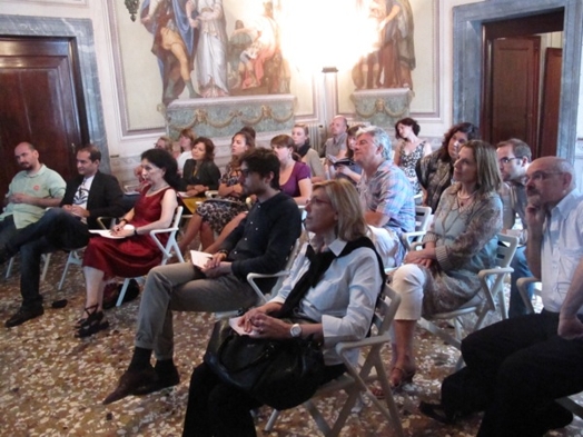 A photo of a group of peoples listening to a lecture, while sitting in a room with frescoes on the walls.