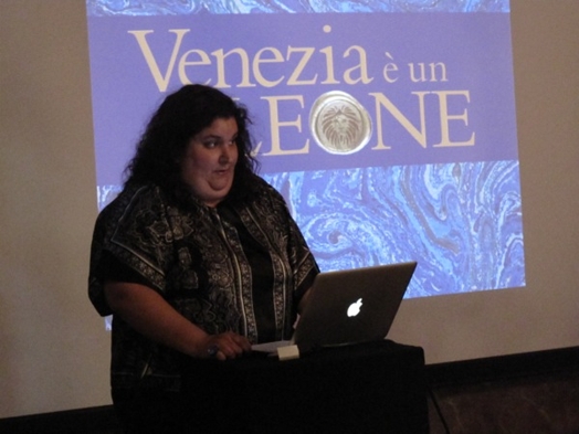 A photo of a woman giving a lecture on Venezia, while having a projected title behind her.