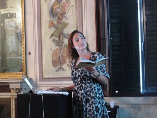 A photo of a person, with a book in her hand and giving a lecture while sitting in a room with frescoes on the walls.