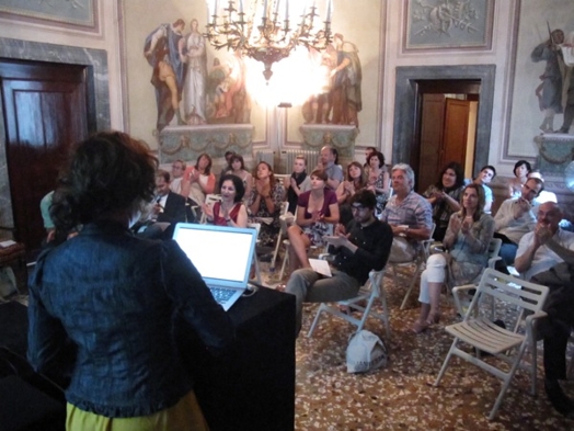 A photo of a group of people listening to a lecture while sitting in a room with frescoes on the walls and a chandelier in the middle.
