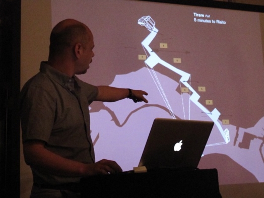 A photo of a person giving a lecture while showing something on the projected screen behind him.
