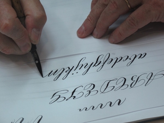 A photo of two hands drawing calligraphic letters on a piece of paper.