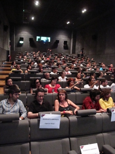 A photo of a group of people sitting in a cinema hall and talking.