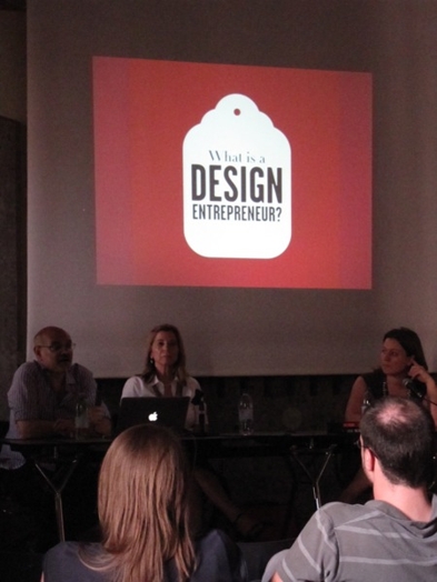 A photo of three persons giving a lecture while behind them is a screen projection of a white logo on a red background with text: What is Design Entrepreneur.