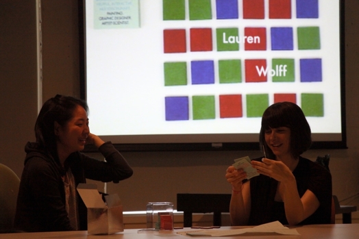 A photo of two women checking some items while in the background is a projected screen with blue, green and red squares.