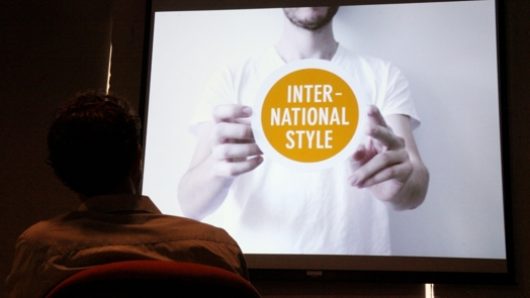 A photo of a man watchin a film projection of another person holding a white and yellow circle that says: International Style.