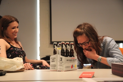 A photo of two laughing women sitting at a desk that has a pack of beers on it.