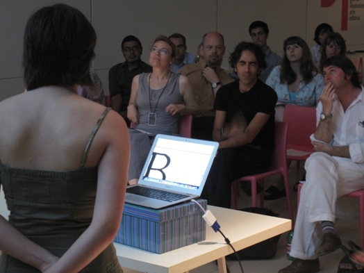 A photo of a group of students listening to a lecture given by a person standing in front of a laptop.