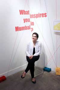 A photo of a woman sitting on a transparent glass stool, in a corner and the phrase What inspires you in Mumbai? is written on the wall.