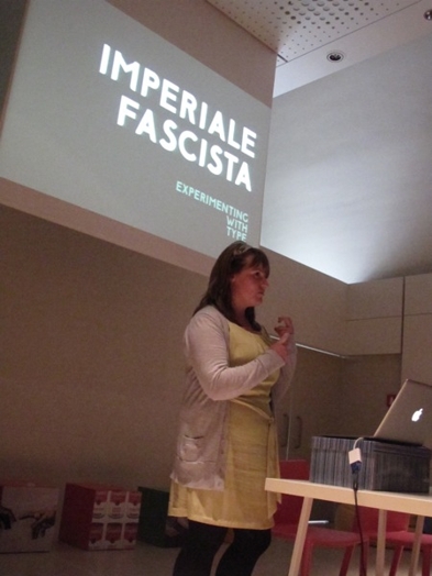A photo of a woman giving a lecture while having behind her the projected white text that says: Imperiale Fascista.