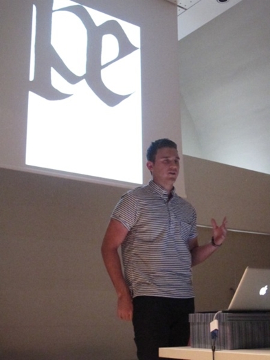 A photo of a man giving a lecture while behind him is a typographic letter on a white projected screen.