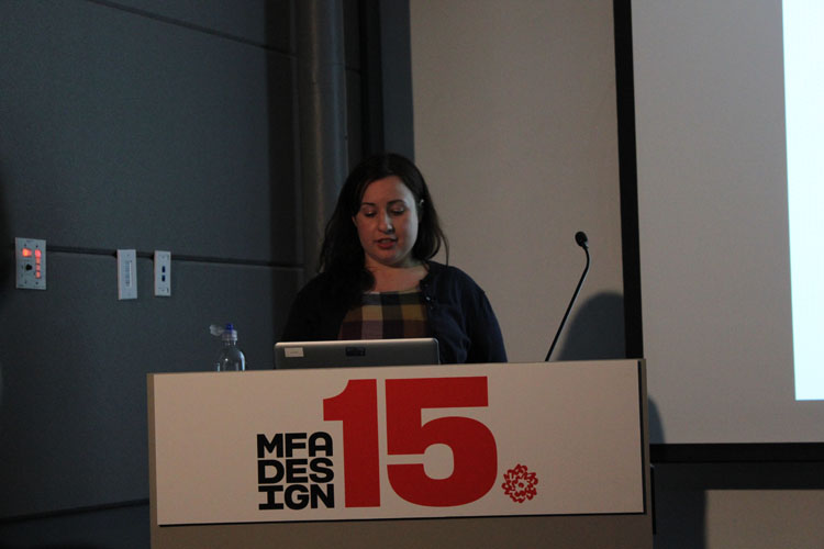 A photo of a woman giving a lecture while sitting at a stand with text MFA DESIGN 15 on it.