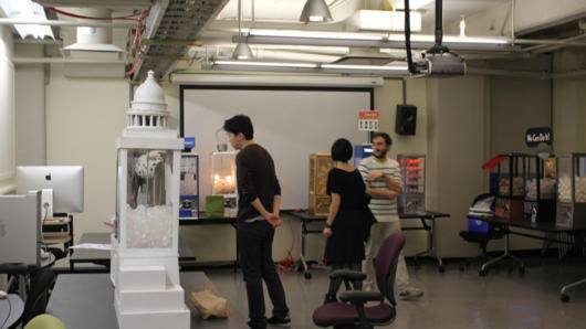 A photo of three students in an art workshop looking at a rubber ball white  vending machine with the capitol on top of it.