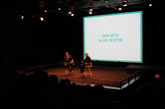 A photo of two people standing on a stage, while near them there is a screen projector with Q and A With Alan Yentob logo.