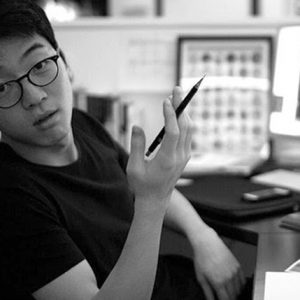 A black and white photo of a man wearing glasses while sitting at a desk and holding a pen.