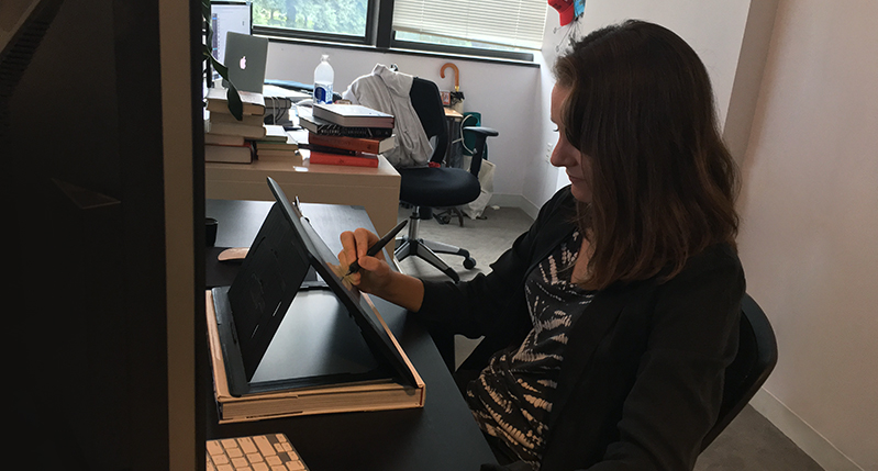 A photo of a woman siting in a room and sketching something on a tablet.