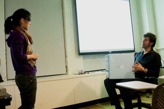 A photo of a man and a woman talking in a classroom while the man holds in his lap an cyan colored box.