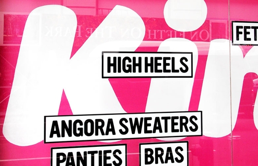 A pink street billboard with text: high heels, angora sweaters, panties, bras.