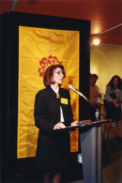 A woman giving a lecture at a stand while having behind her an orange cloth banner with the red SVA logo on it.