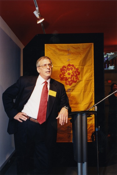 A man giving a lecture at a stand while having behind her an orange cloth banner with the red SVA logo on it.