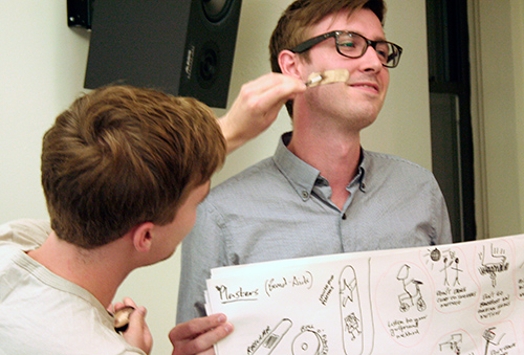 A man wearing glasses and holding a sketch board being painted on the face by another man.