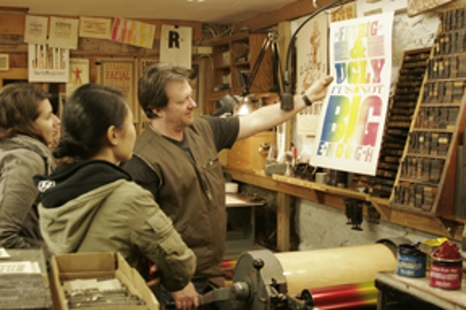 A photo of a man ant two women looking at a freshly printed poster while sitting in a typographic room.