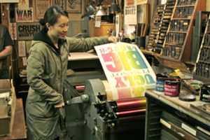 A photo of a woman that pulls a fresh printed, rainbow collared paper from a typography press machine.