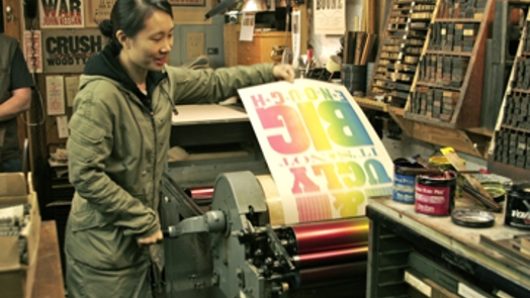 A photo of a woman that pulls a fresh printed, rainbow collared paper from a typography press machine.