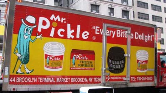A commercial on a side of a truck depicting a cyan personified picke, some jars. and a barrel. On a red and yellow banner the text says: Mr. Pickle Every Bite a Delight.