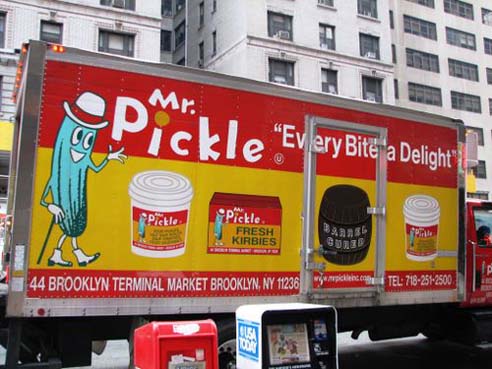 A commercial on a side of a truck depicting a cyan personified picke, some jars. and a barrel. On a red and yellow banner the text says: Mr. Pickle Every Bite a Delight.