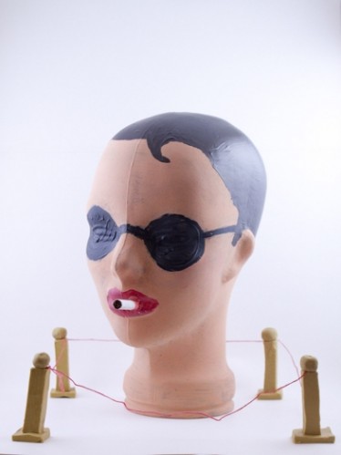 A woman's head made from plastic material, with swimming cap and glasses, smoking a cigarette and enclosed by a single thread fence and four pillars.