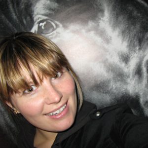 A photo of a woman wearing a black coat in front of a painting that shows  what looks like an eye.