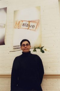A photo of a man wearing glasses and holding a white rose in his mouth, while standing near a white wall with posters, on of which has the text slave on it.