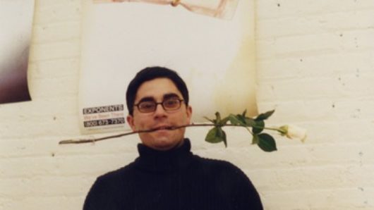 A photo of a man wearing glasses and holding a white rose in his mouth, while standing near a white wall with posters, on of which has the text slave on it.