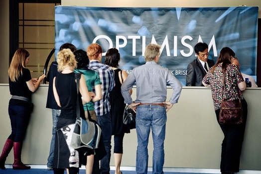 A group of people standing and waiting at the reception desk while behind it is a blue banner with text: Optimism.