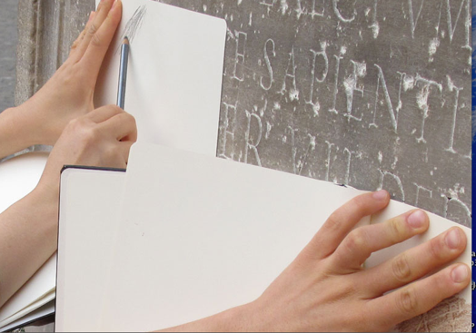 A photo of a pair of hands holding pieces of paper against an engraved wall and copying the writings with a pencil.