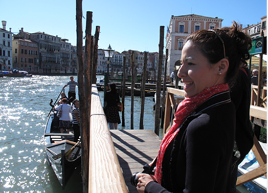 A photo of a woman standing at a dock near a gondola on one of Venice's channels.