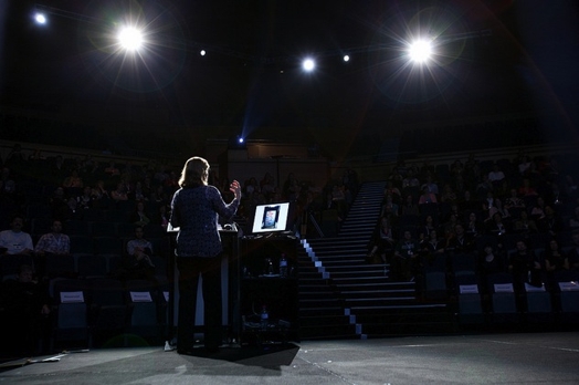 A photo of a woman giving a lecture while standing on a stage.