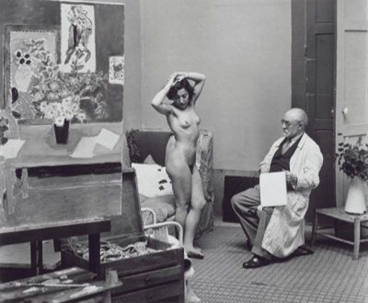 A black and white photo of an old man sitting on a chair and holding a sketchbook while looking at a naked woman in an art studio.