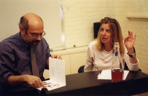 A photo of two people talking to each other while one of them is checking a sketchbook.